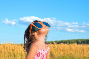 Young girl wearing a sun dress and looking up to the sky through oversized sunglasses while standing in a wheat field.