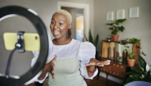 Smiling young African female influencer standing in her living room at home and talking during a video using a smart phone