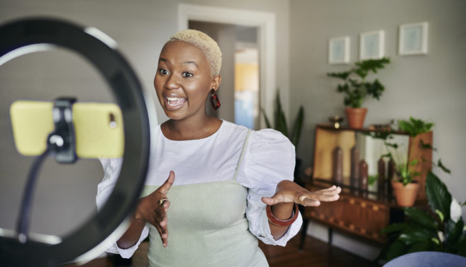 Smiling young African female influencer standing in her living room at home and talking during a video using a smart phone
