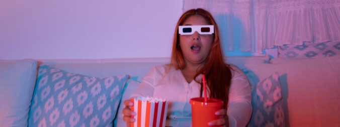 Woman with 3D glasses with popcorn and drink sitting on a couch.